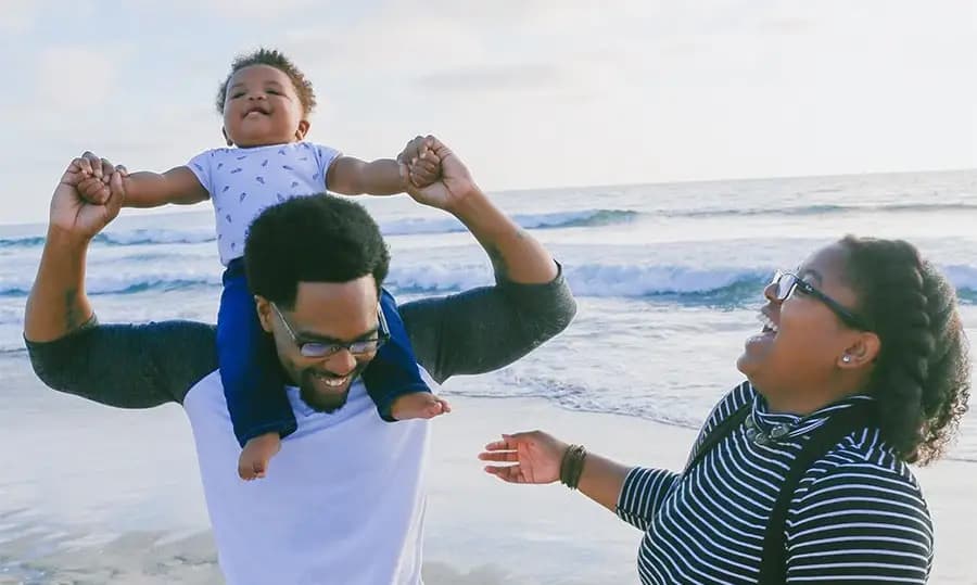 Man carries baby on his shoulders and laughs with wife on beach