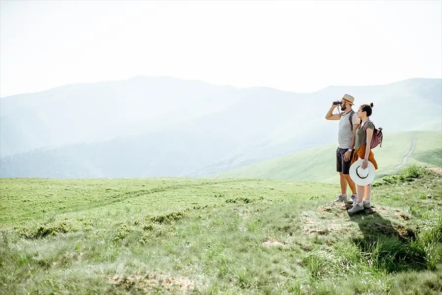 Two people stand on top of a hill, looking beyond with binoculars