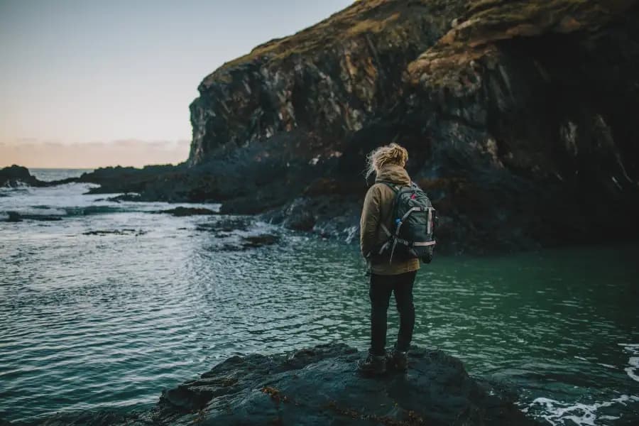 Woman in a backpack stands on a rock looking out at the ocean