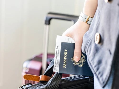 Woman's hand holding US passport and vaccination record with suitcase in background.